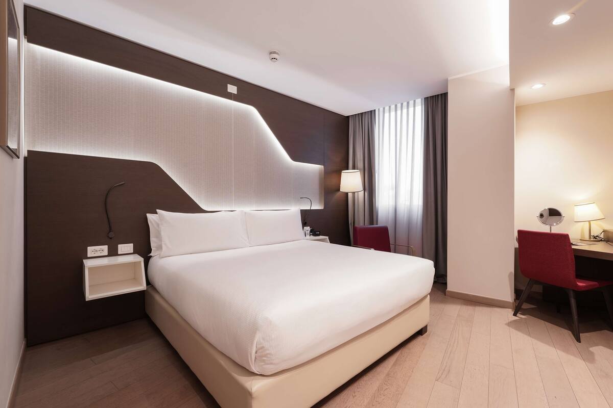 Ein modernes Hotelzimmer mit einem großen Bett, einem Schreibtisch mit Stuhl, einem Nachttisch und einer Lampe.
