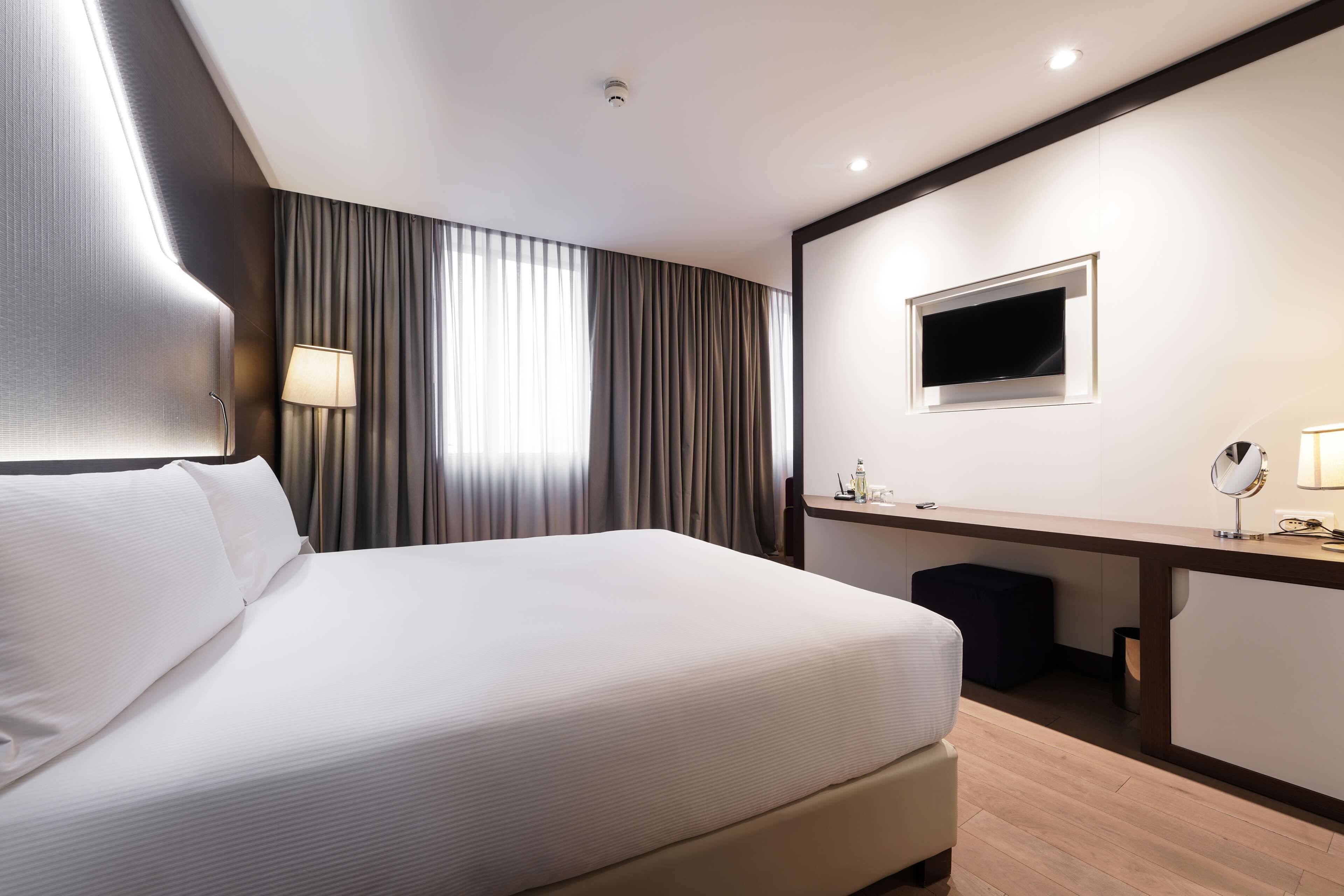 A modern hotel room with a large bed, a desk with a flat-screen TV, a lamp, and a window with curtains.