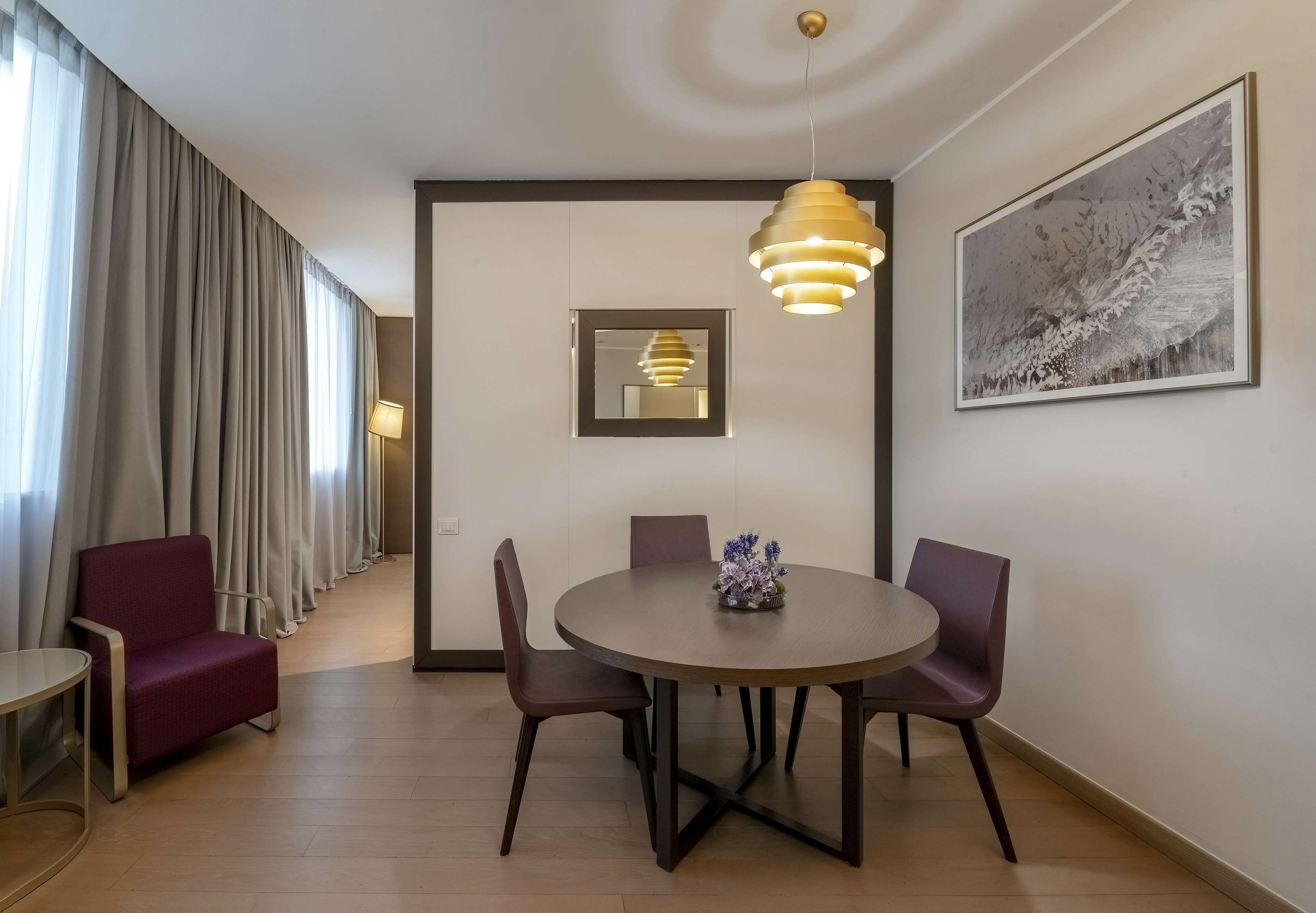 Un comedor moderno con una mesa redonda, cuatro sillas, una lámpara colgante, una obra de arte enmarcada en la pared y una ventana con cortinas.