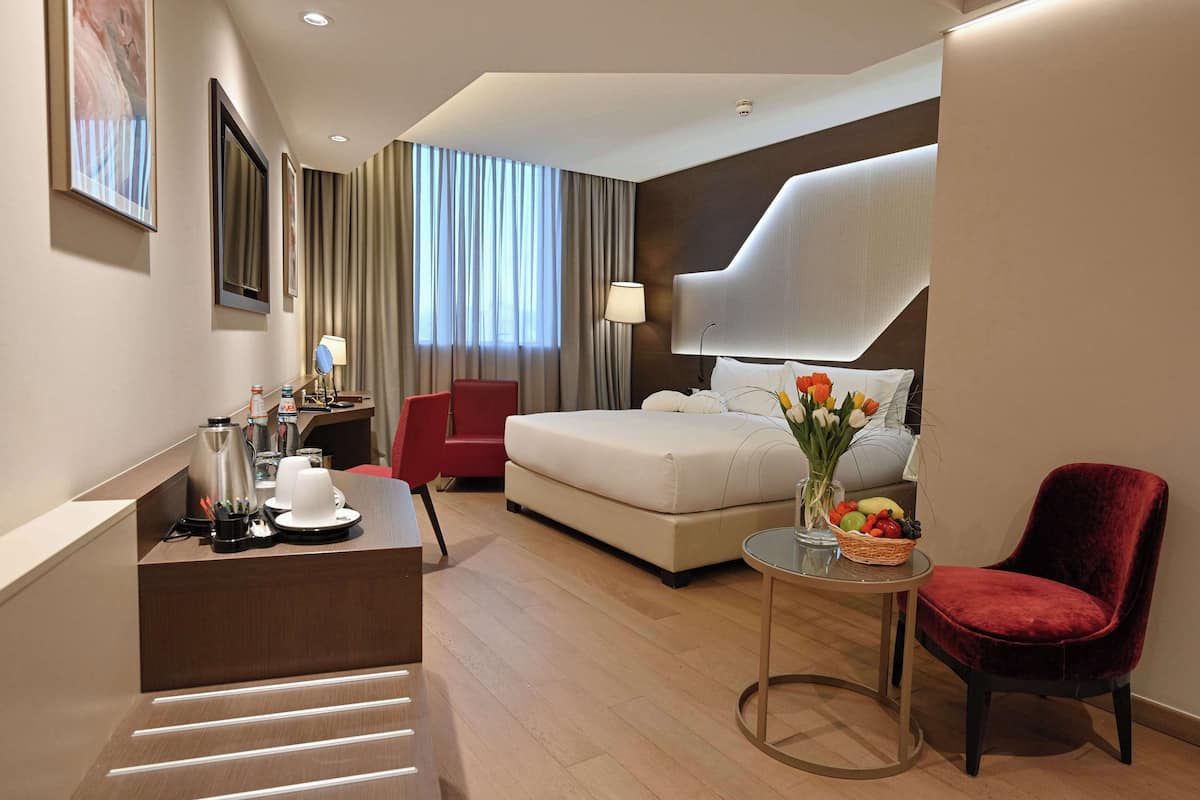 Ein Hotelzimmer mit einem Bett, einem Schreibtisch mit Kaffeemaschine, einem roten Sessel, einem kleinen Tisch mit Blumenvase und einem großen Fenster mit Vorhängen.