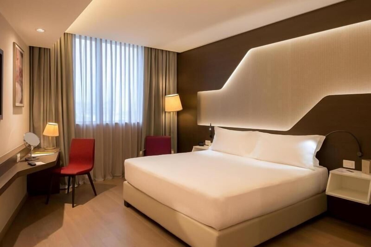A modern hotel room with a large bed, a desk with a chair, a nightstand, and a window with curtains.