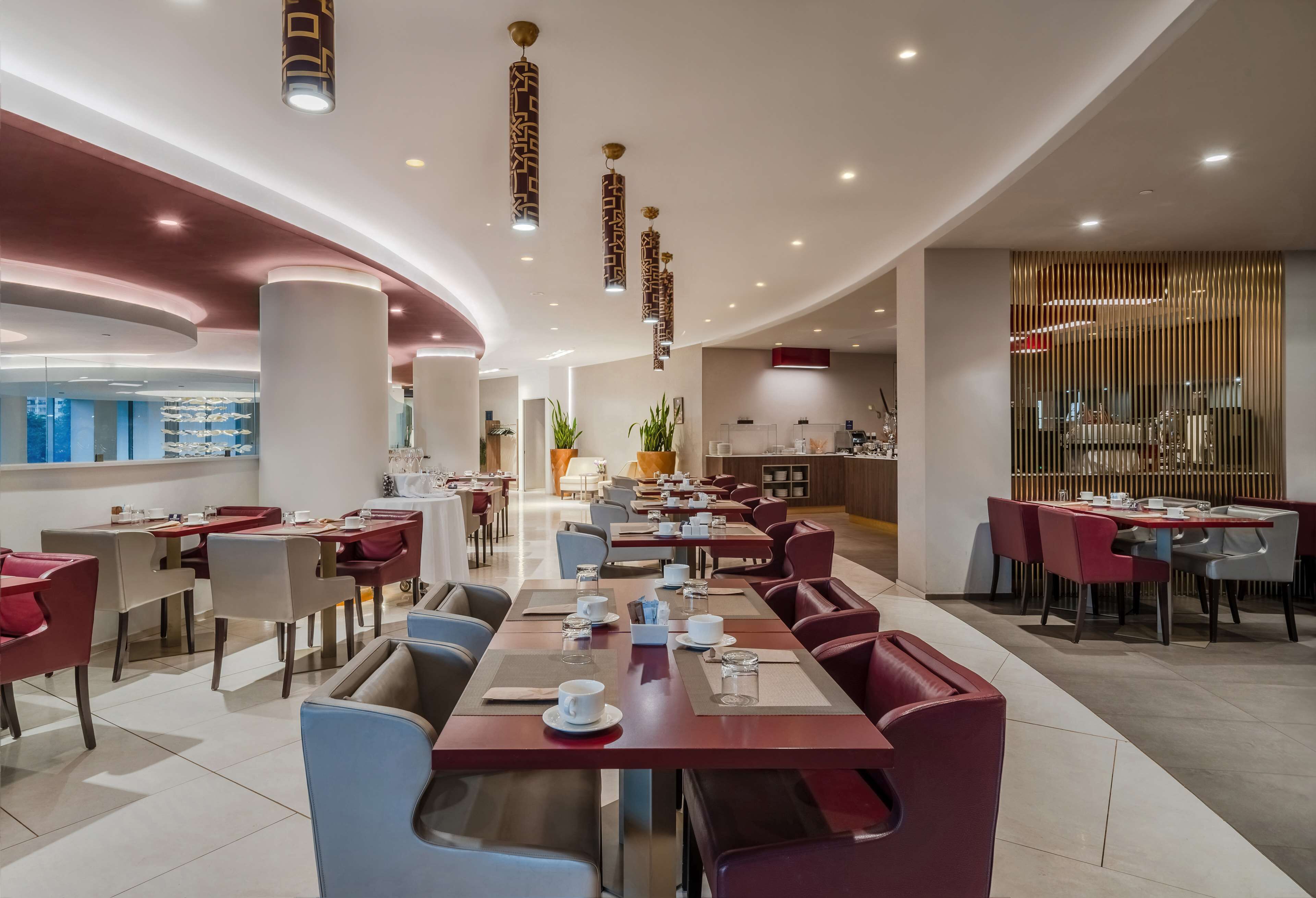 A modern dining area with red and beige seating, a kitchen in the background, and pendant lighting.