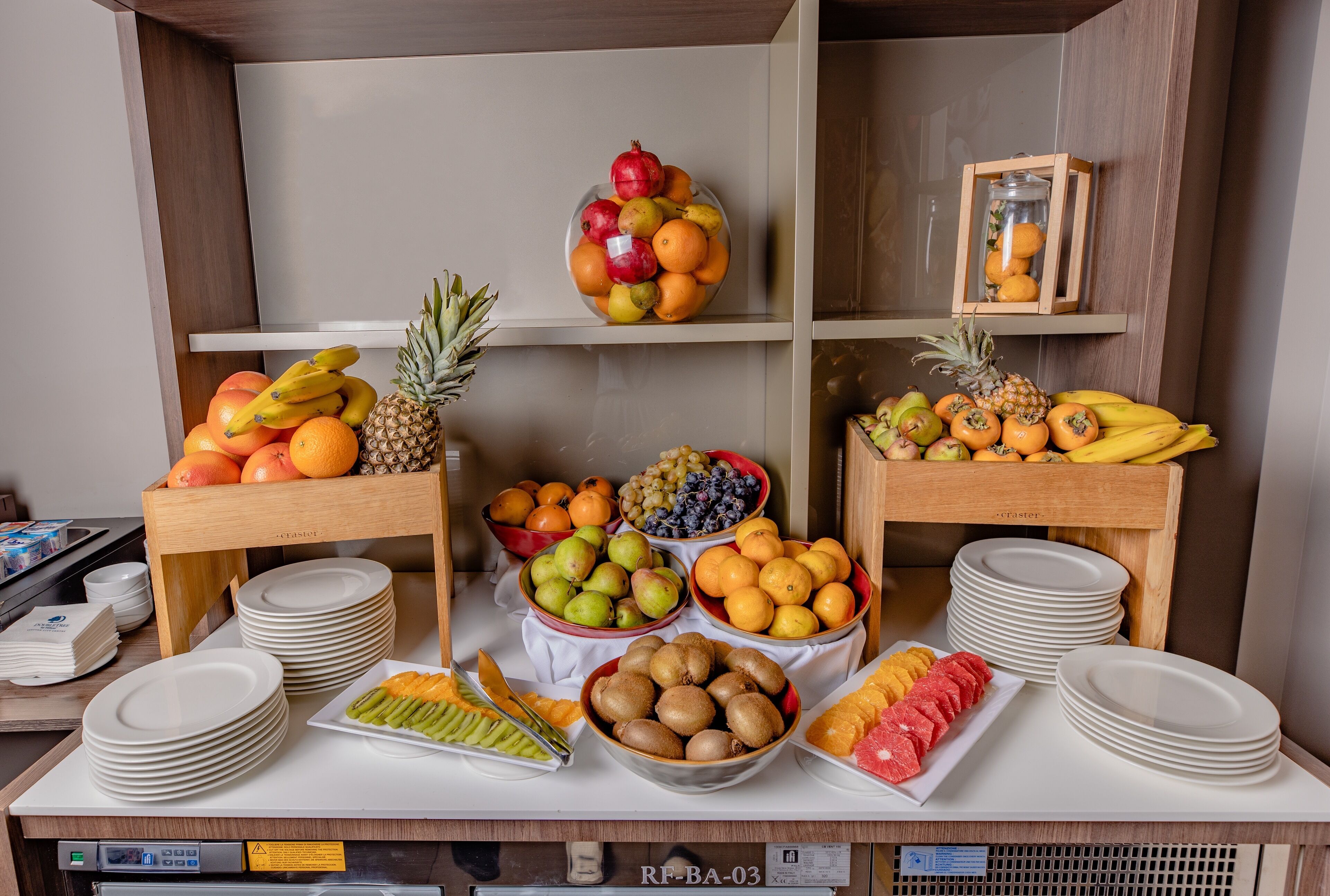 Een buffettafel met diverse soorten fruit en groenten, zoals bananen, ananassen, sinaasappels en kippen, uitgestald op een witte tafel met gestapelde witte borden.