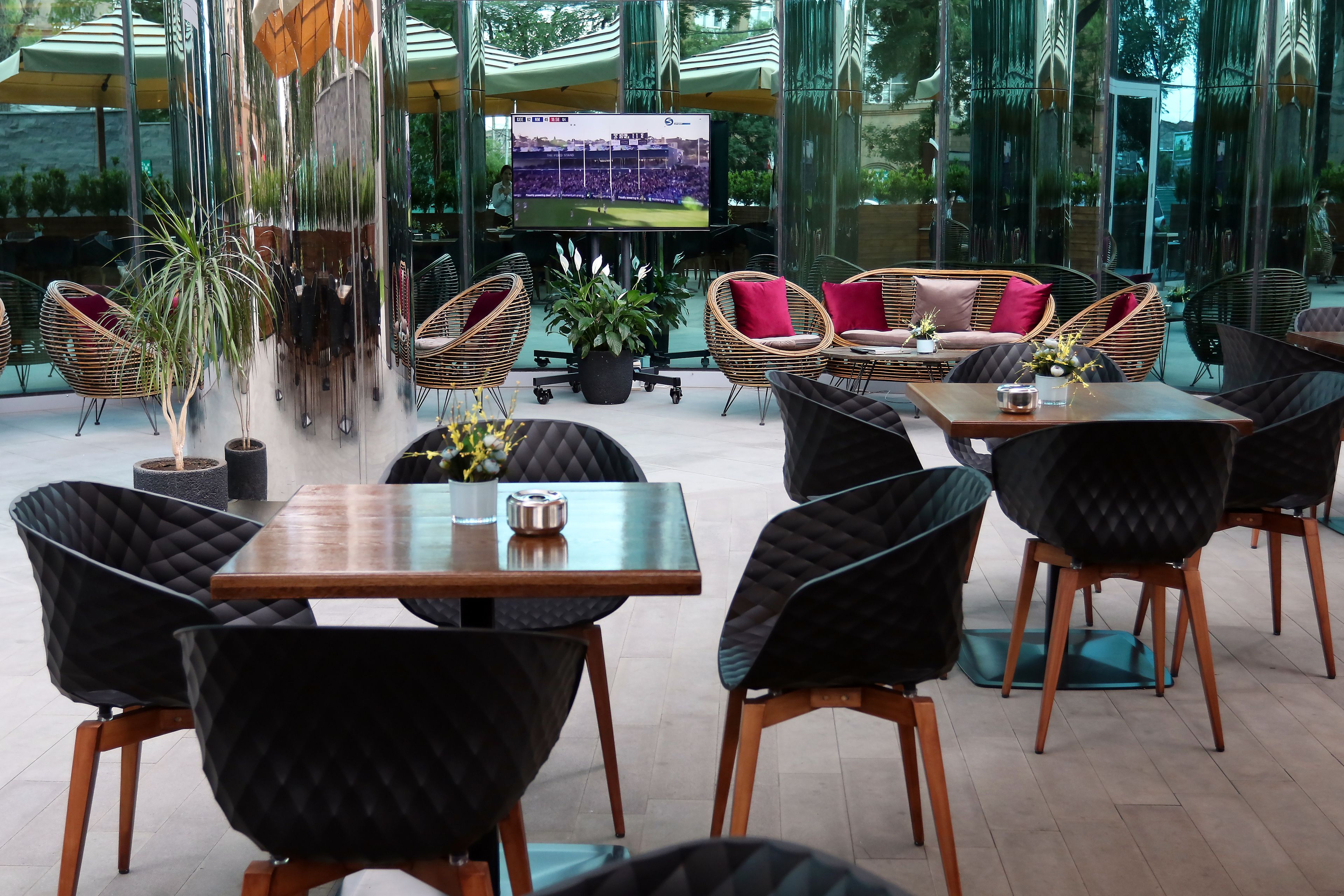 A modern outdoor seating area with wicker chairs and tables, a large TV screen displaying a sports event, and large glass windows offering views of the surrounding area.