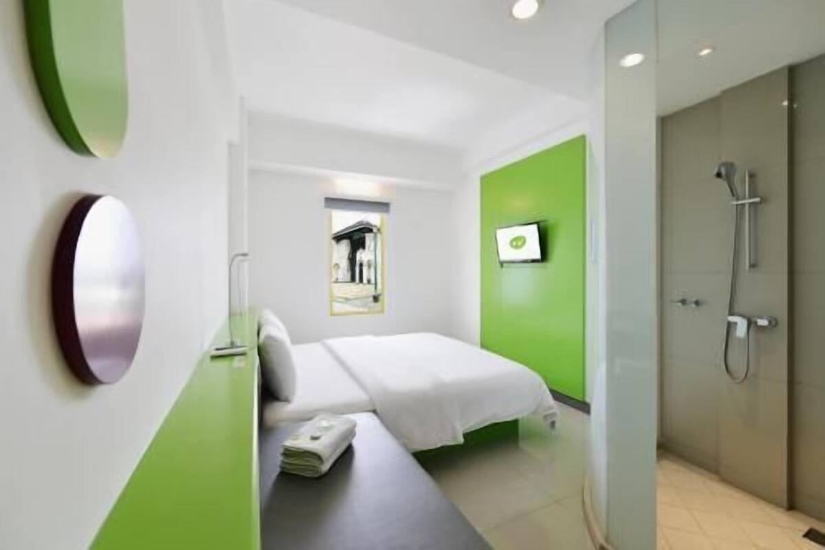 Una habitación de hotel moderna con cama, mesita de noche, pared de acento verde, ducha y televisor montado.
