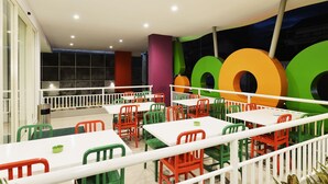 A brightly colored indoor dining area with white tables and chairs, a white railing, and a mural with large circular shapes.