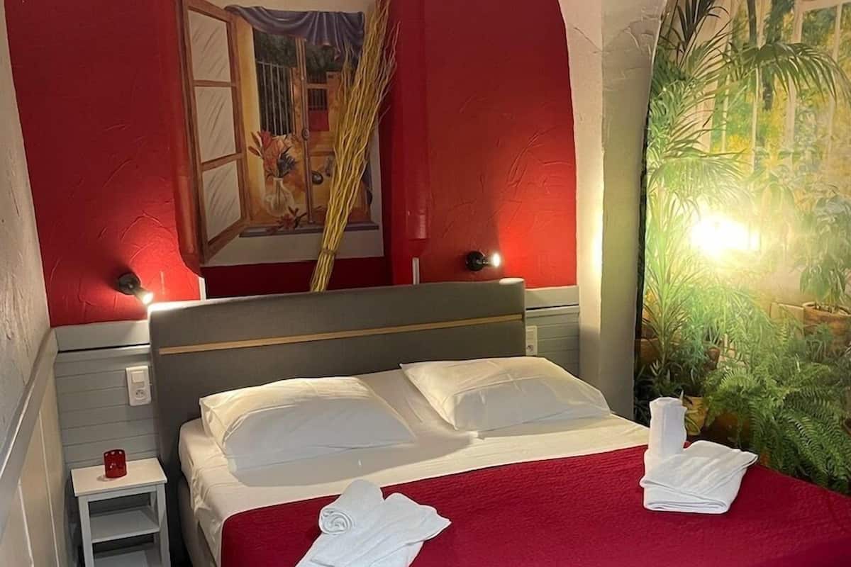 A neatly made bed with white linens and a red blanket, a small bedside table with a red candle, and a large window with a view of a tropical garden.