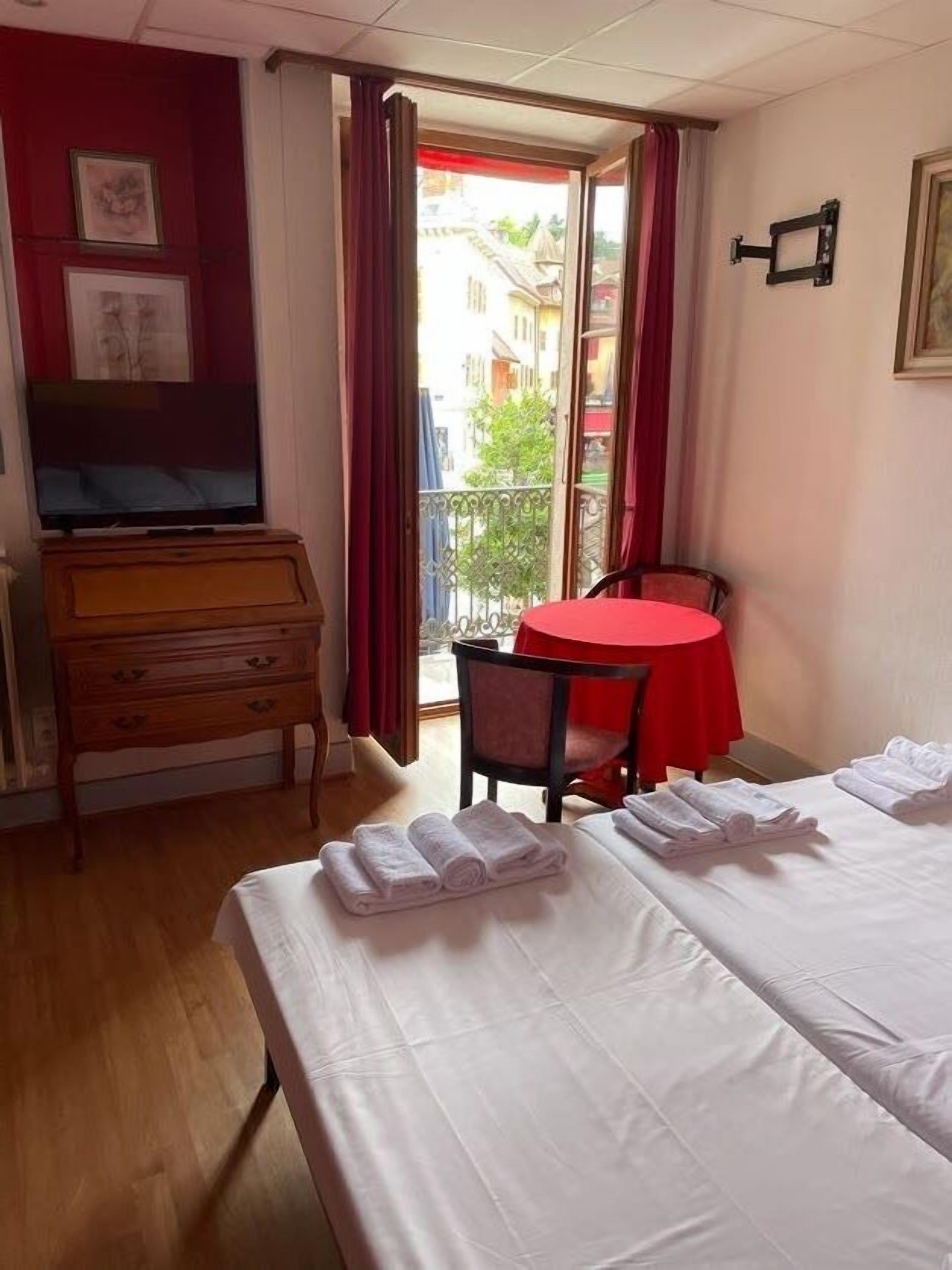 A hotel room with a bed, a TV, a wooden dresser, a red table, and a balcony with a view of buildings.