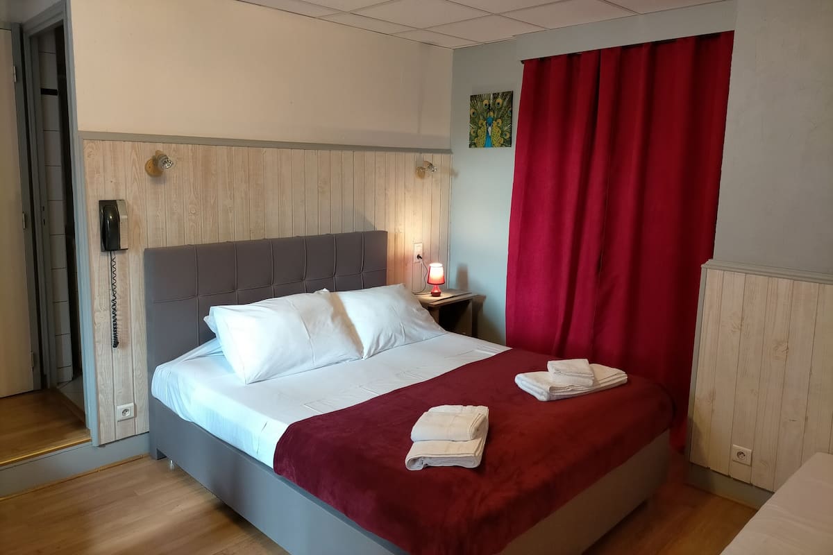 A hotel room with a bed, a red curtain, a nightstand with a lamp, and a wooden panel wall.