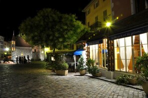 Une place pavée, avec un bâtiment historique doté d’une tour horlogée, un restaurant proposant des tables en plein air et des plantes en pot.