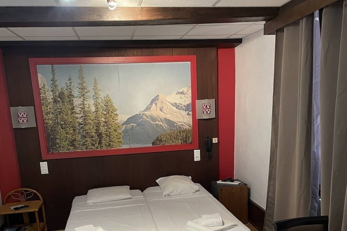 A hotel room with a double bed, a wooden bench, a small table, and a large window with a mountain view.
