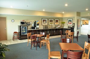 A hotel kitchen with wooden tables and chairs, a microwave, coffee machine, and a refrigerator. - Brookstone Lodge & Suites (Algona)