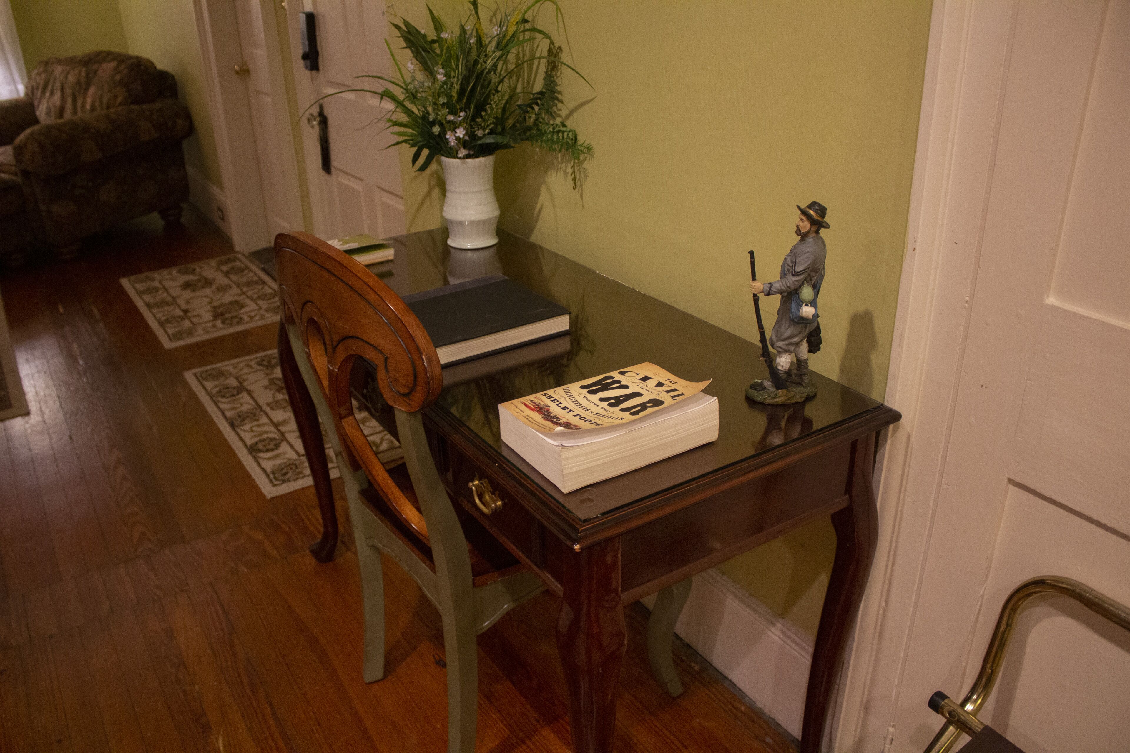 A wooden table with a book titled "War" and a figurine. A chair is positioned at the table, and a potted plant is on the wall.