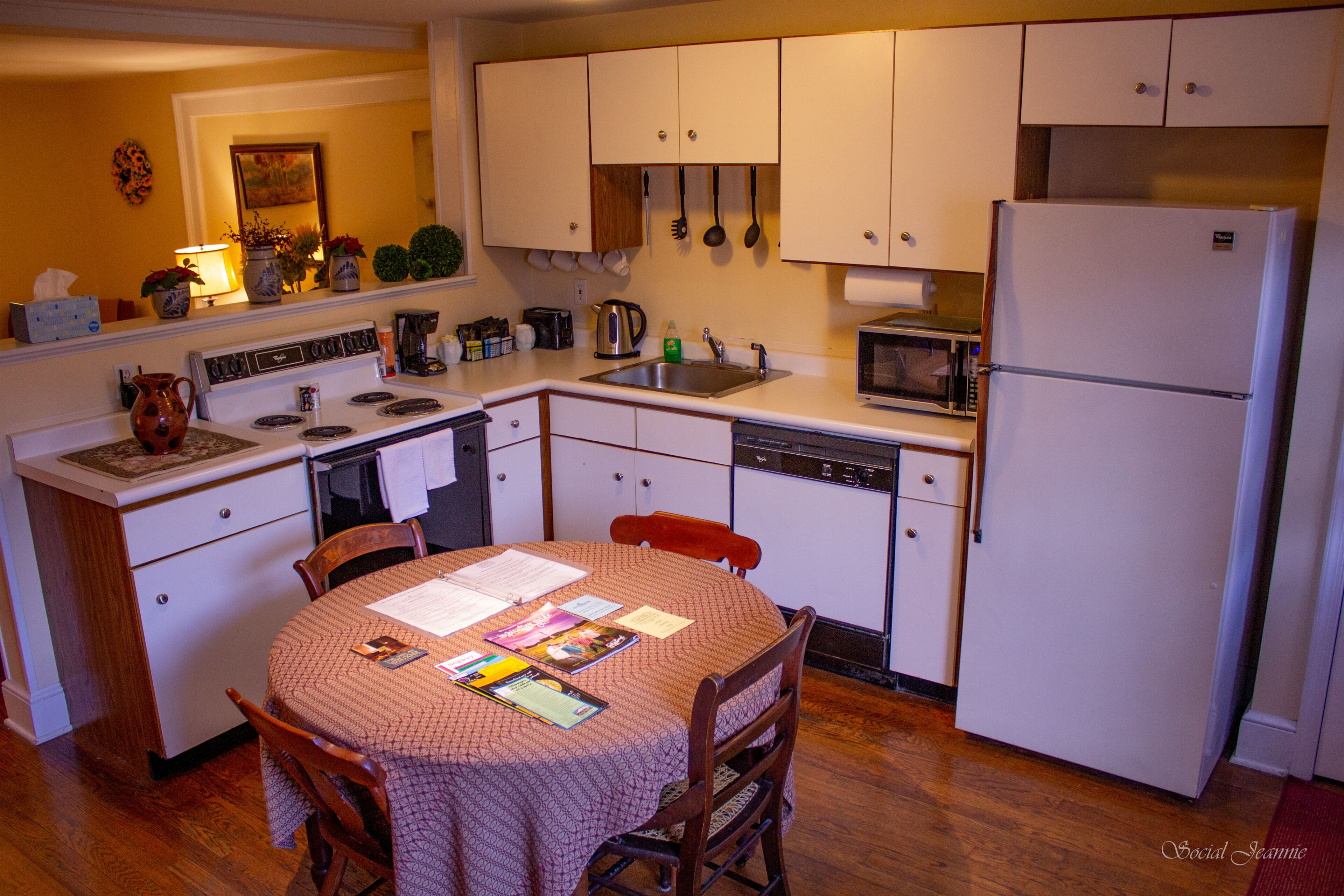 Une cuisine compacte avec des meubles blancs, une cuisinière, un évier, un four à micro-ondes et un réfrigérateur. On y trouve une table à manger avec des chaises et un vase posé sur le comptoir.