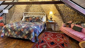 A cozy attic bedroom with a wooden ceiling, a brick wall, a bed with a colorful quilt, a nightstand with a lamp, and a pink sofa. - Inn at Lincoln Square (Gettysburg)