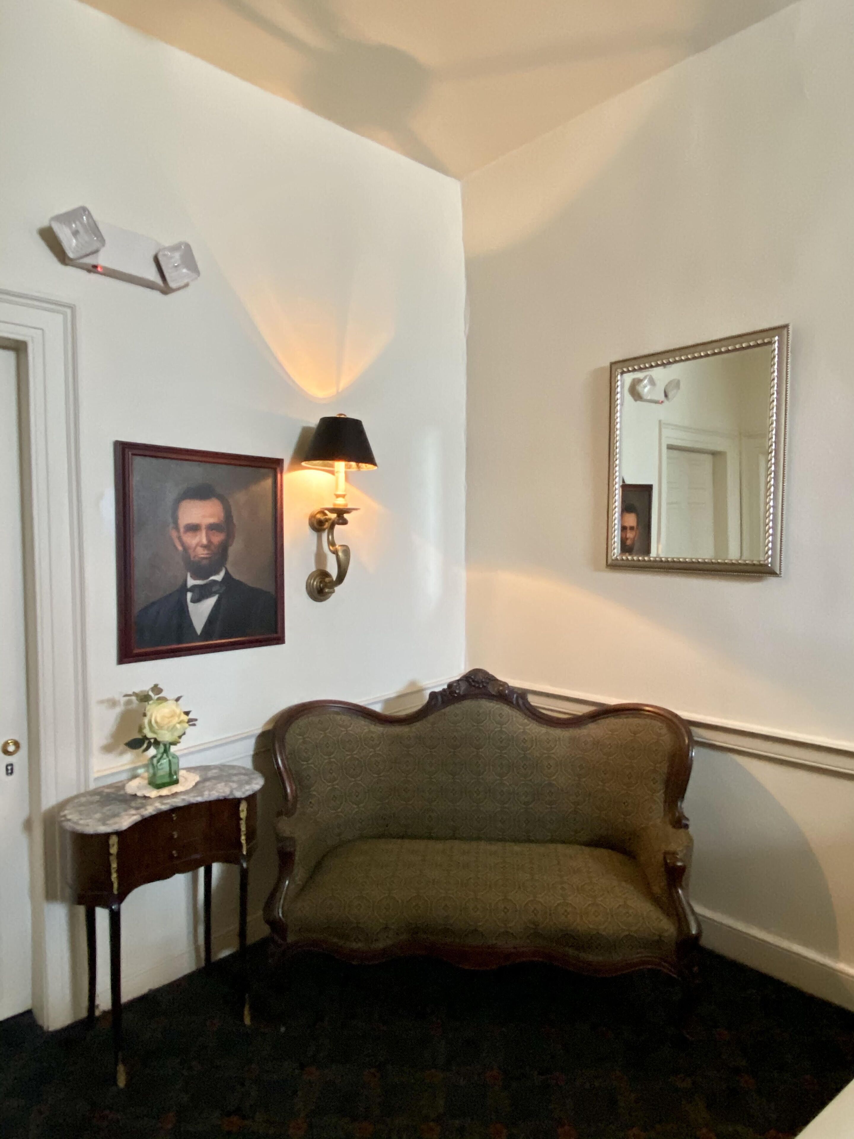 Ein Zimmer mit einem gerahmten Porträt, einer Wandlampe, einem Spiegel, einem kleinen Tisch mit einer Vase und einem gemusterten Sofa.