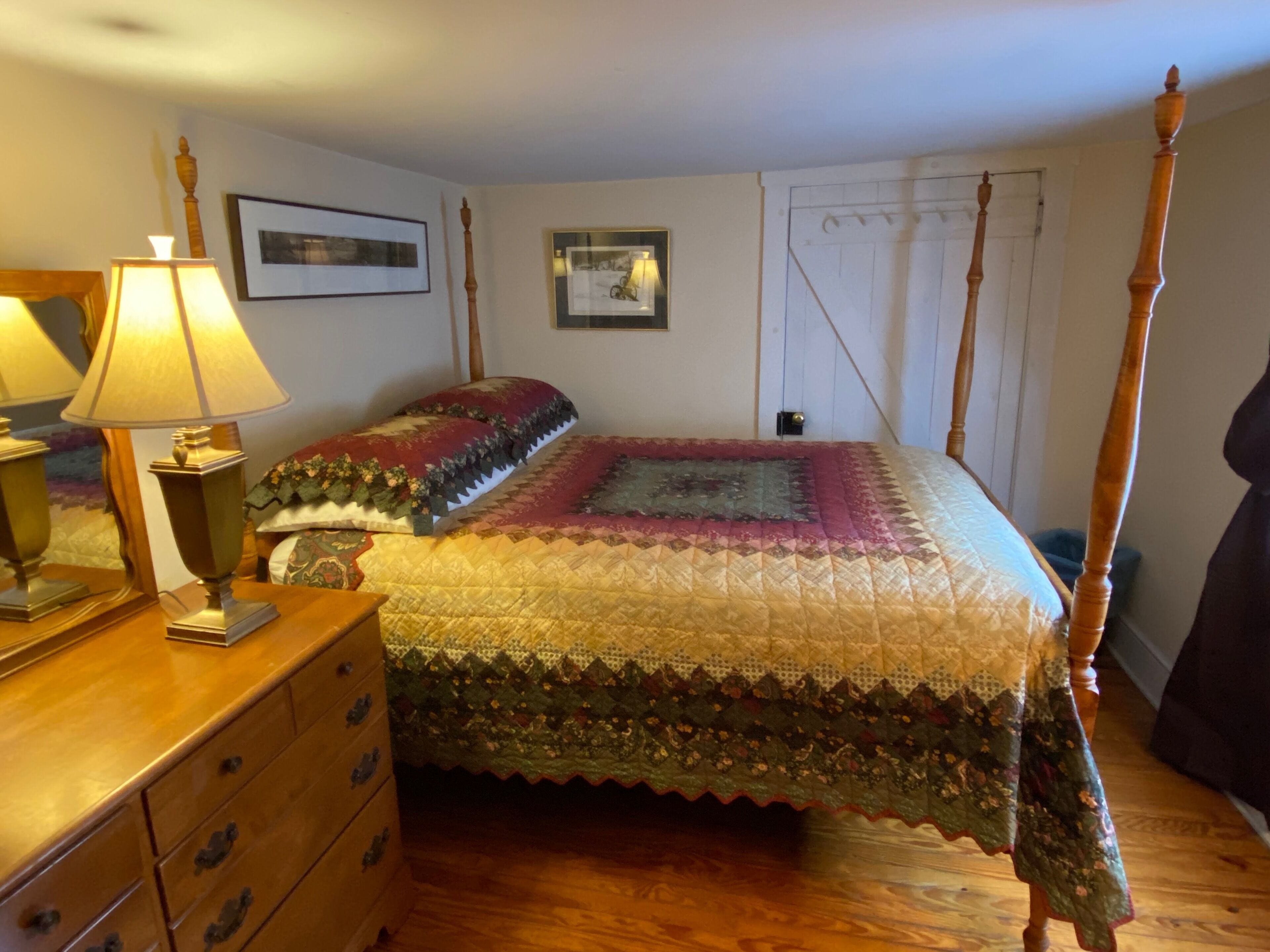 Uma cama de dossel com colcha, um aparador de madeira com gavetas, um abajur e quadros nas paredes.