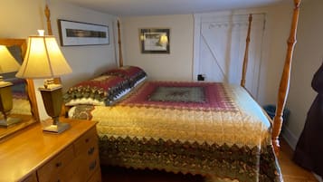 Uma cama de dossel com colcha, um aparador de madeira com gavetas, um abajur e quadros nas paredes.
