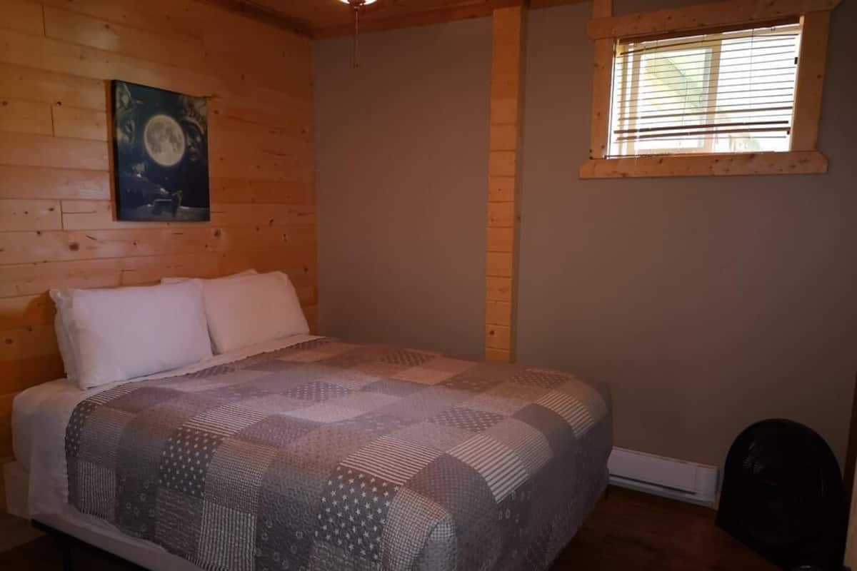 A bedroom with a wooden floor, a bed with a patterned blanket, a night sky painting on the wall, and a window with blinds.
