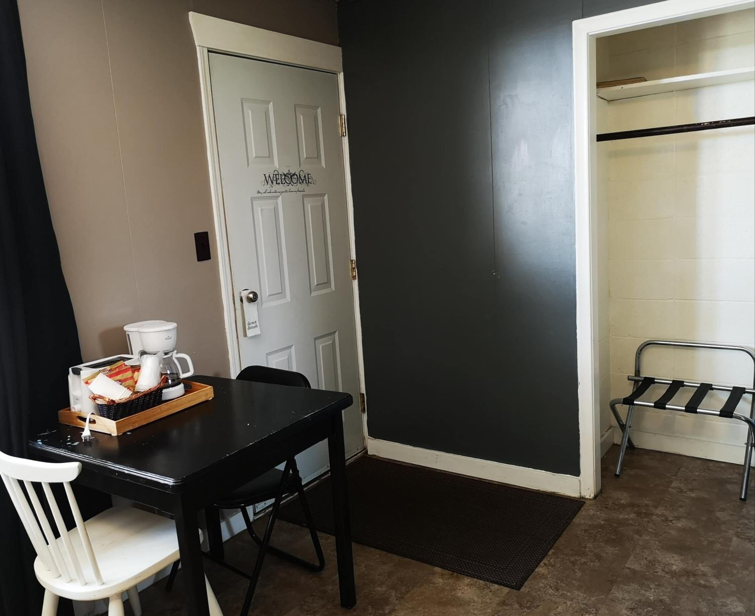 Una habitación con una mesa negra, una silla blanca y una cafetera sobre la mesa. Hay una puerta blanca con pomo y un letrero que dice “Welcome”. 