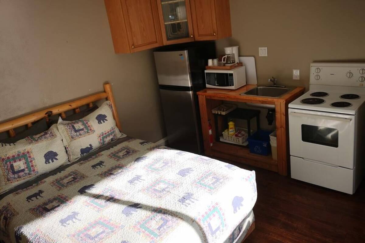 A compact kitchen with wooden cabinets, a refrigerator, a microwave, a stove, and a bed with a patterned blanket.