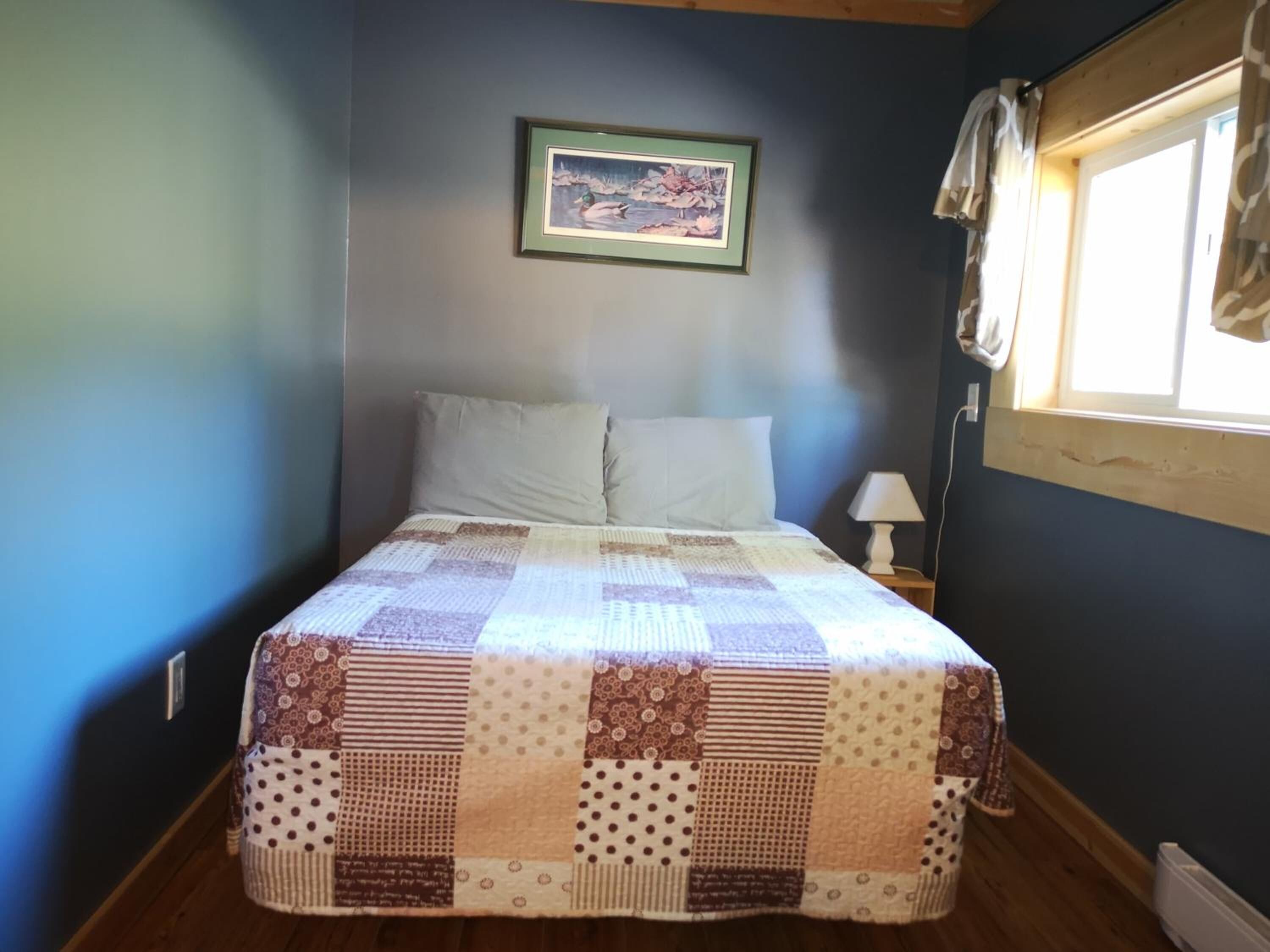 Un dormitorio con cama, una mesita de noche con lámpara, un cuadro enmarcado en la pared y una ventana con cortinas.