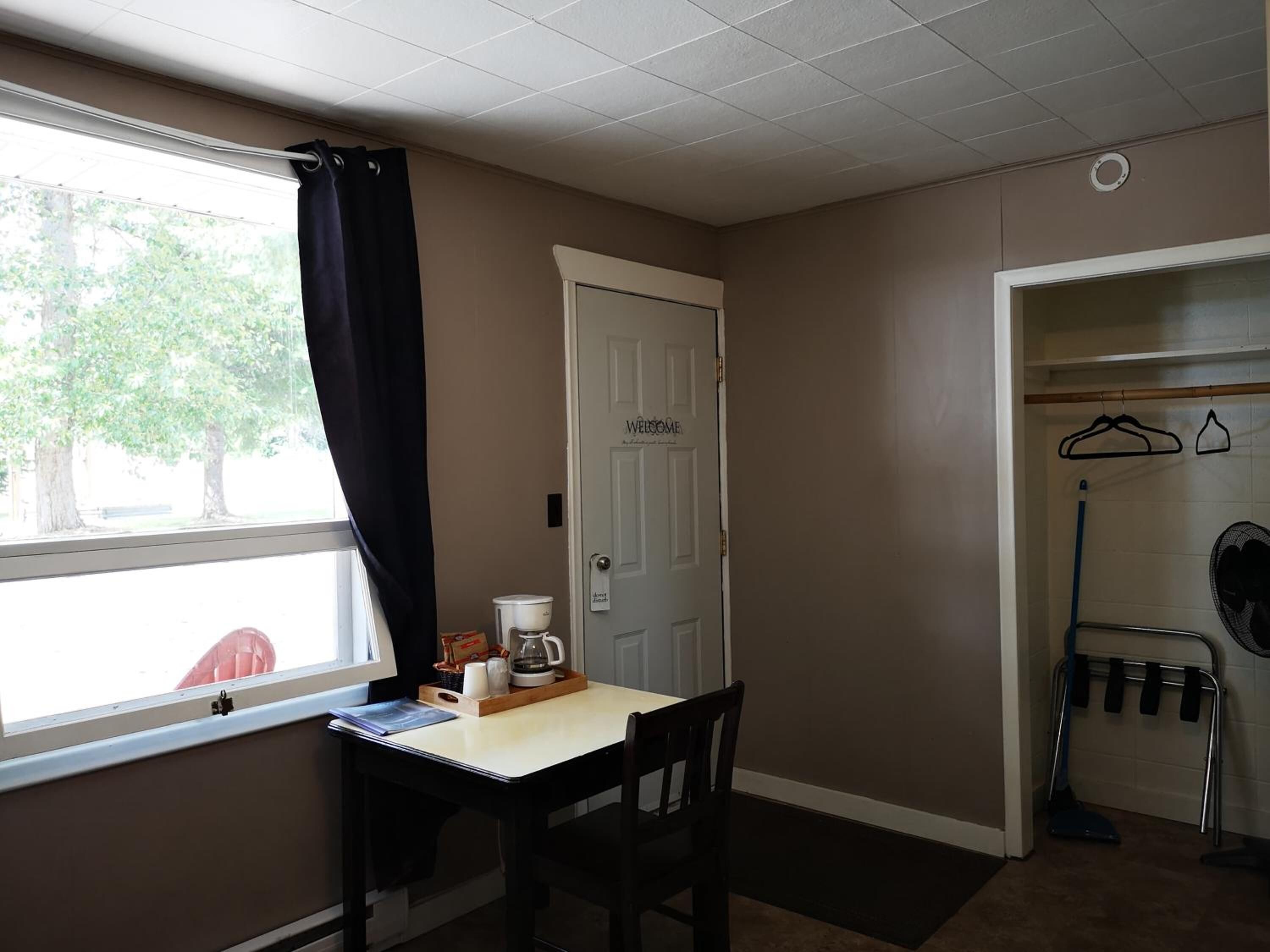 A small room with a window, a door, a table with a coffee maker, and a chair.