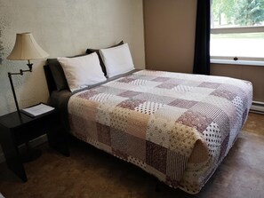 A neatly made bed with a patterned quilt, a bedside lamp, and a window with a view of greenery. - Overlook Inn & Cabins (Clearwater)
