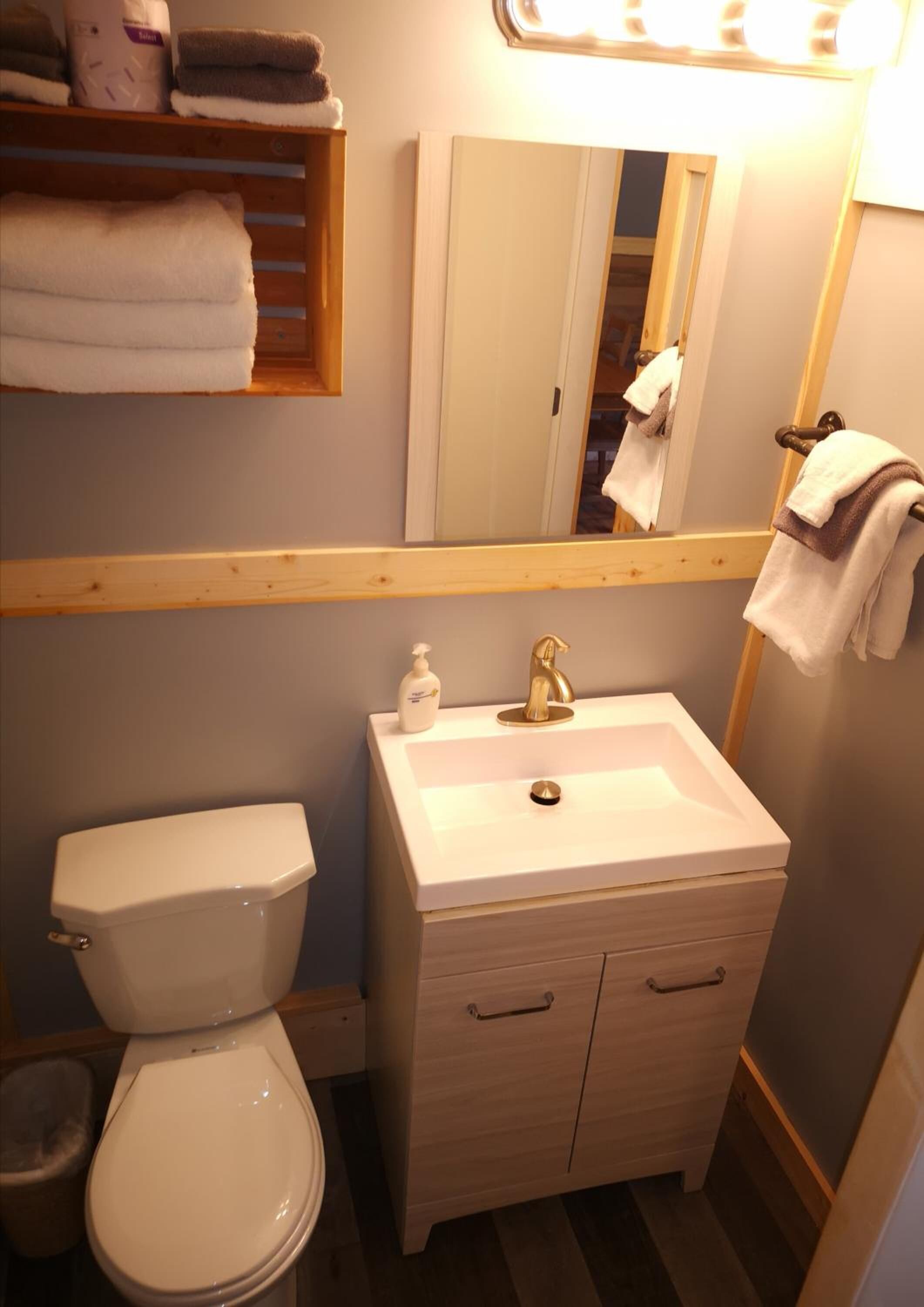 A bathroom with a white toilet, a sink with a gold faucet, and a mirror above the sink. There are towels on a shelf above the sink and a towel hanging on the wall.