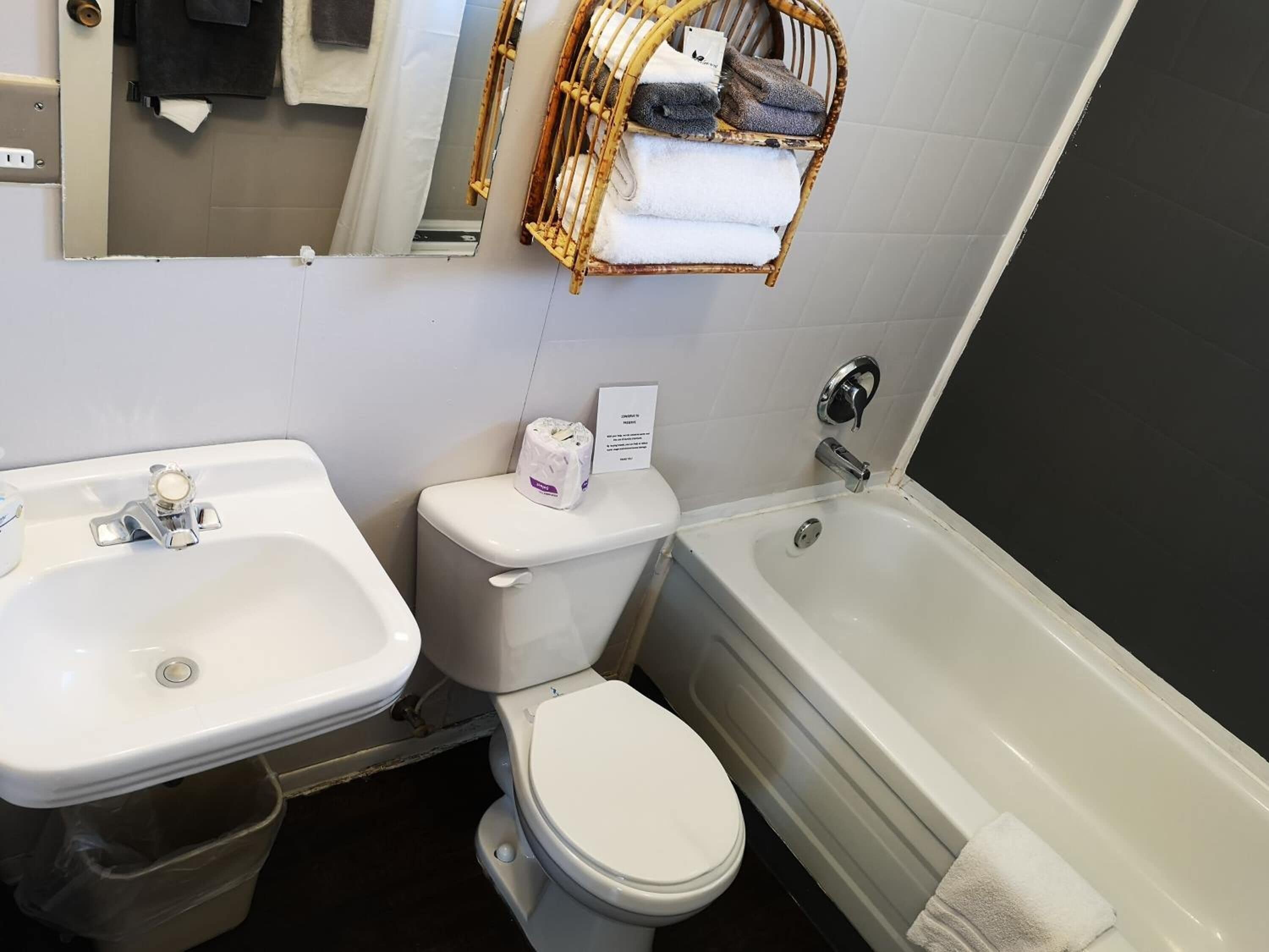 Un baño con lavamanos, inodoro y bañera. Hay un perchero sobre el inodoro y un cubo de basura junto al lavamanos.