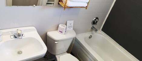 Un baño con lavamanos, inodoro y bañera. Hay un perchero sobre el inodoro y un cubo de basura junto al lavamanos.