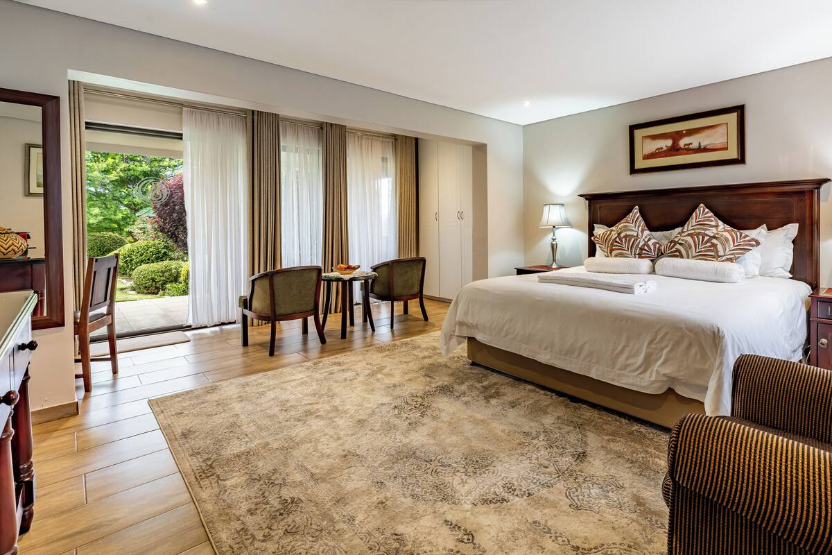 A hotel room with a large bed, two chairs, a desk, and a view of greenery through the window.