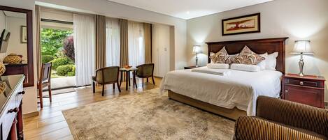 A hotel room with a large bed, two chairs, a desk, and a view of greenery through the window.