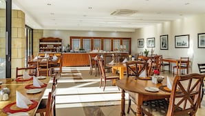 A spacious dining area with multiple wooden tables and chairs, a central bar area with a counter, and a kitchen visible in the background. - Botleng Guest House (Maseru)