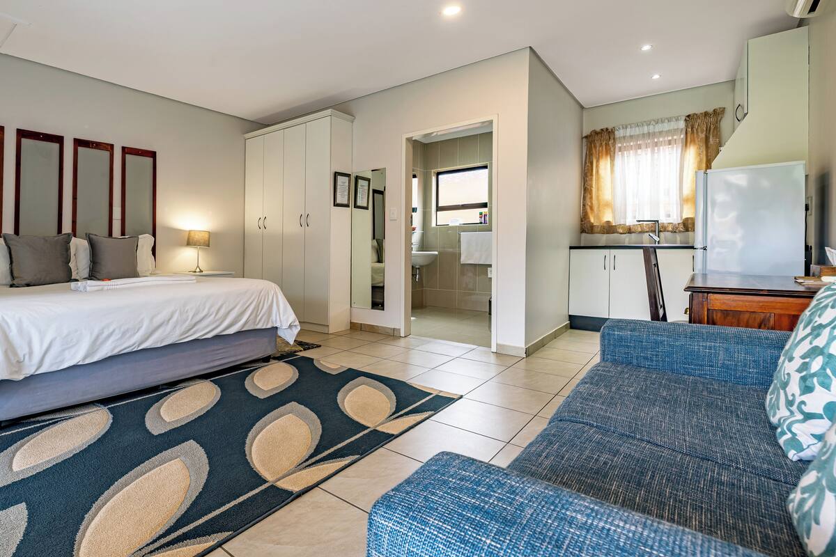 A modern hotel room with a neatly made bed, a sofa, a dining area, and a bathroom visible through an open door.