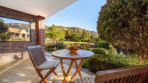 A patio with a round wooden table, two striped chairs, and a view of a garden and mountains. - Botleng Guest House (Maseru)