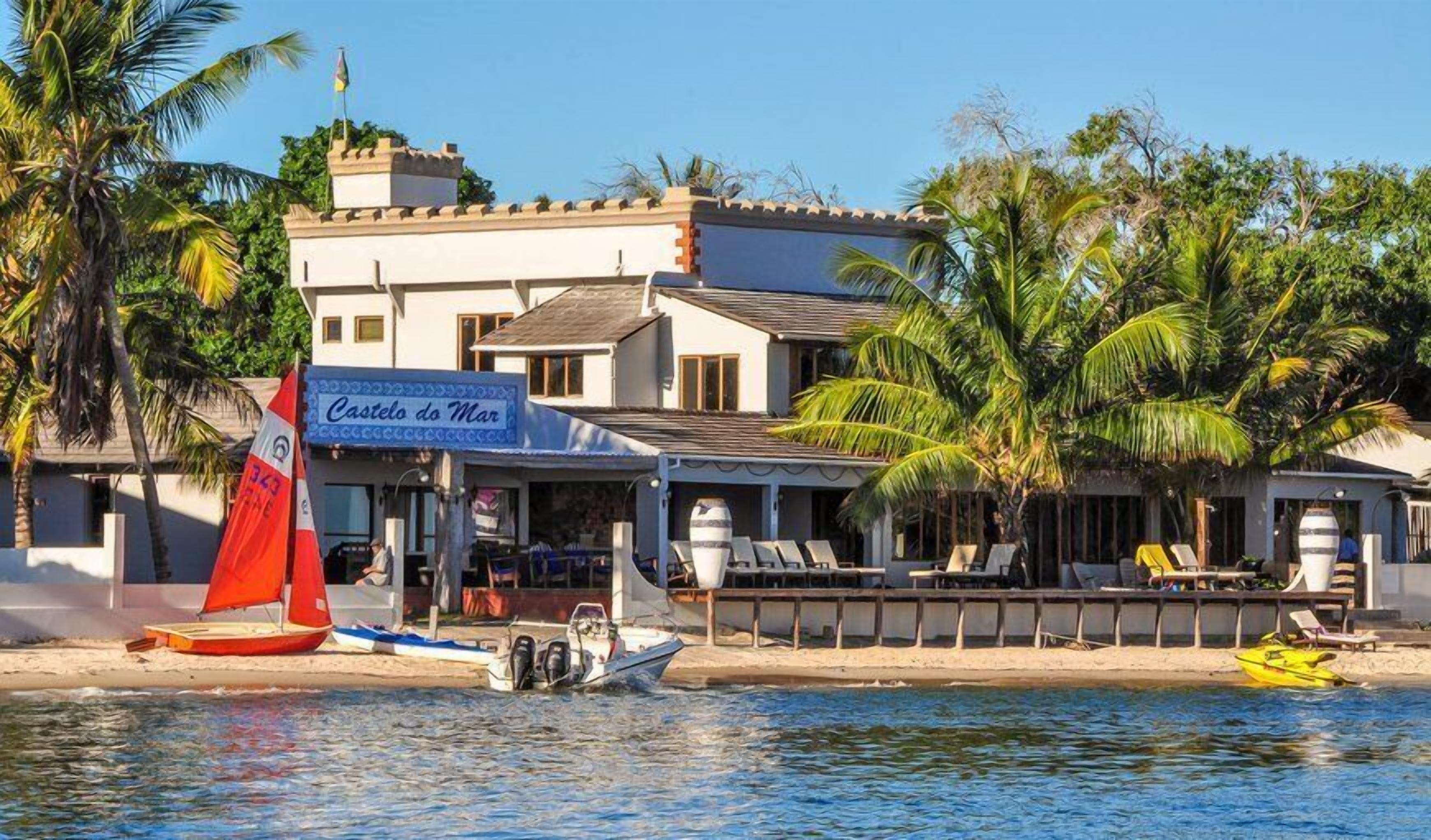 A beachfront property with a sign reading "Castelo do Mar," featuring a boat, lounge chairs, and palm trees.