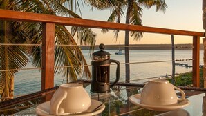A table with two white cups and a black coffee pot, set against a backdrop of palm trees and a view of the sea.