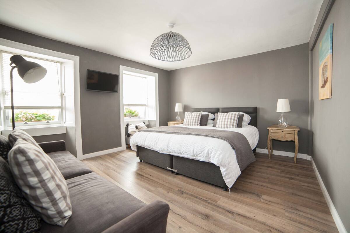 A bedroom with a large bed, a sofa, a TV, and a window with blinds.