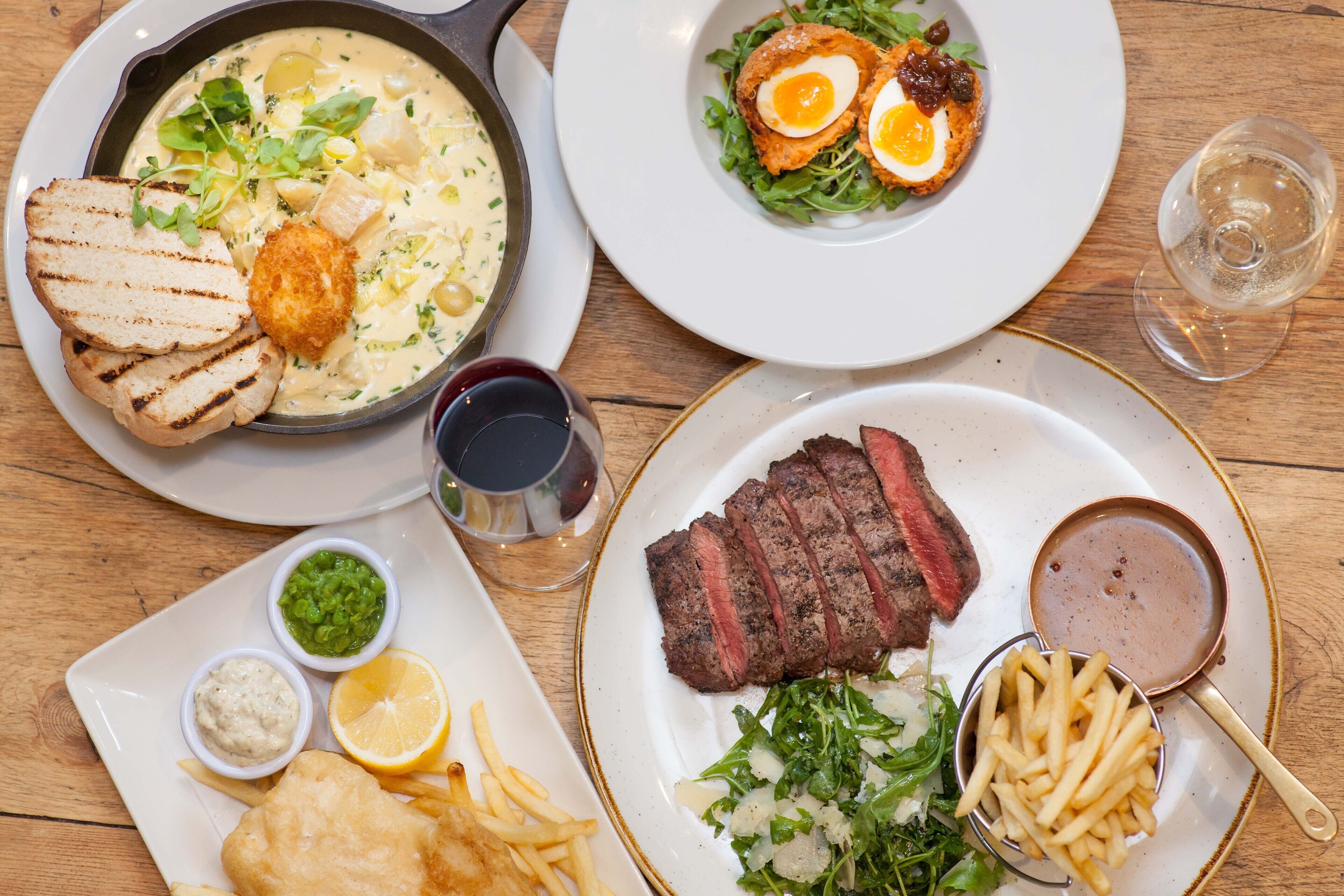 Variedad de platillos, incluyendo bistec, pescado con papas fritas y una sopa cremosa, servidos en platos blancos con detalles dorados.