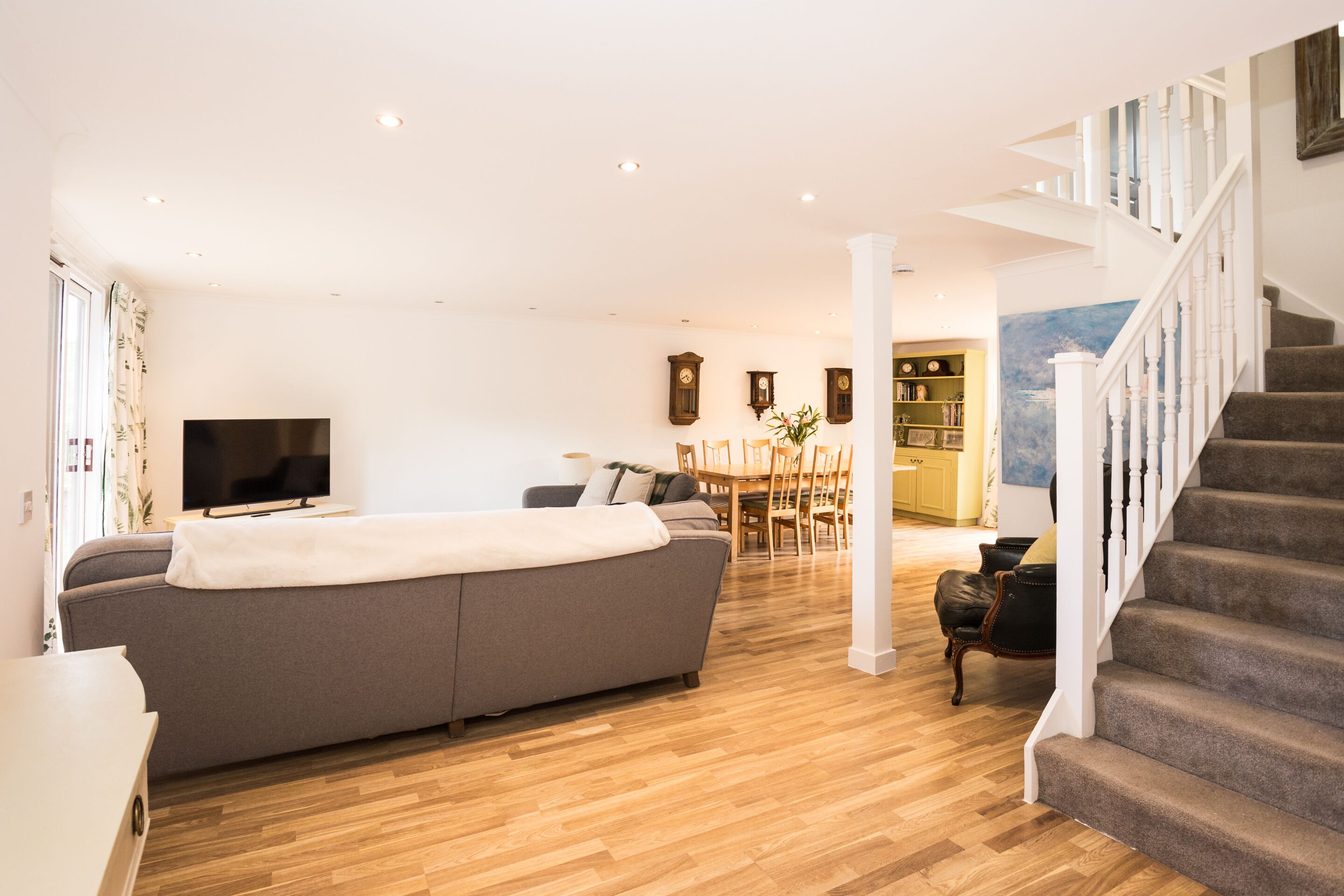 Un salon moderne avec un grand canapé gris, un plancher en bois, un téléviseur à écran plat, un coin repas avec des chaises et une table, et un escalier avec des rampes blanches.