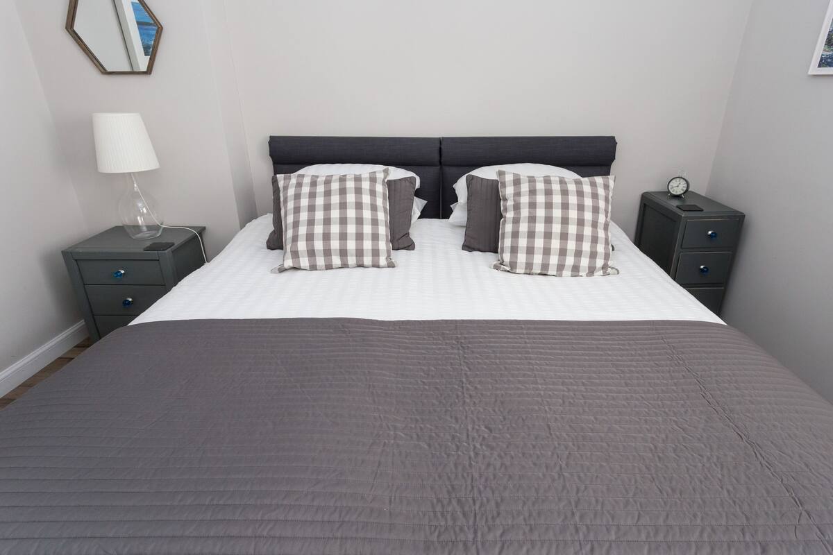 A neatly made bed with a checkered pillow set, a bedside lamp, and a nightstand with a clock.