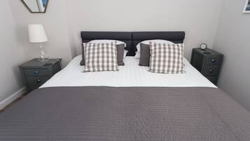 A neatly made bed with a checkered pillow set, a bedside lamp, and a nightstand with a clock.