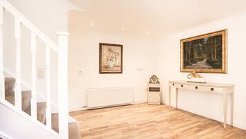 A room with a wooden floor, a white staircase, a radiator, a console table with a clock, and two framed pictures on the wall.