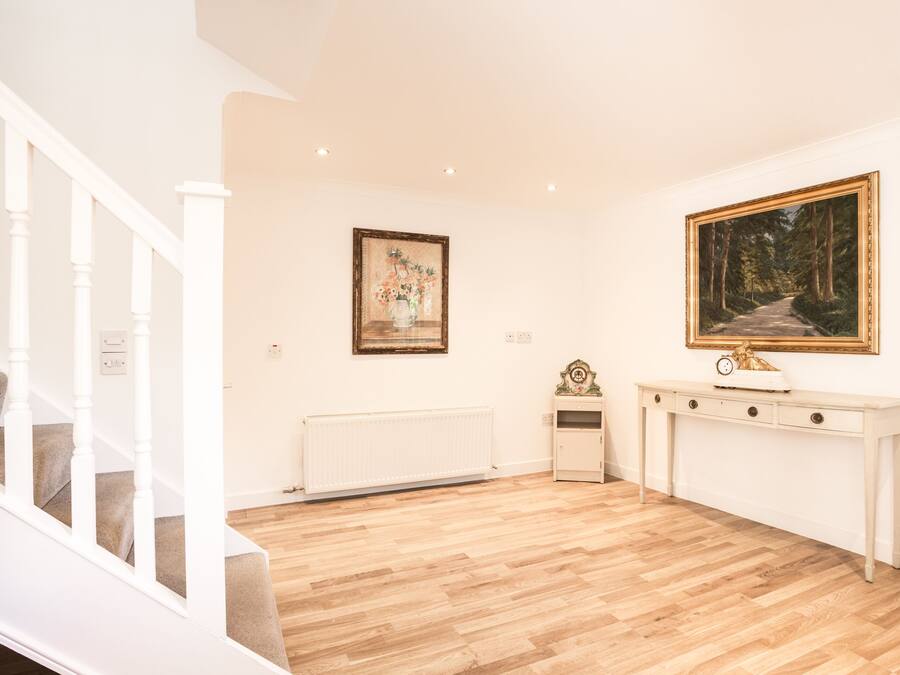 A room with a wooden floor, a white staircase, a radiator, a console table with a clock, and two framed pictures on the wall.