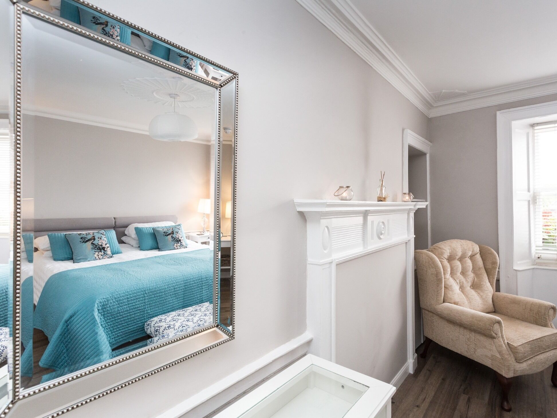 A bedroom with a large mirror, a bed with blue bedding, a white fireplace, and a beige armchair.