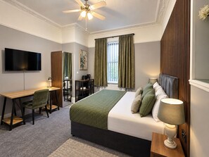 A hotel room with a bed, a desk, a chair, a television, a window with curtains, and a ceiling fan. - Ashfield Hotel (Huddersfield)