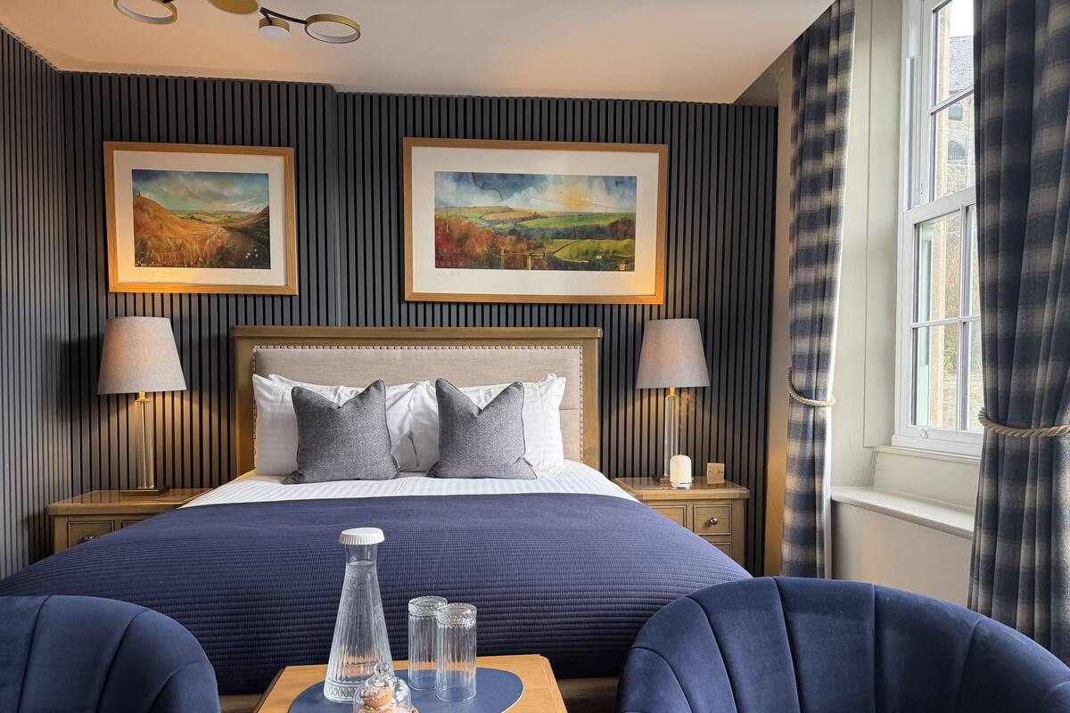 A hotel room with a large bed, two bedside lamps, a nightstand with a lamp, a table with a glass pitcher and sugar bowl, and two chairs.