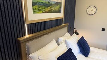 Una cama bien hecha con almohadas blancas y azules, un cuadro de paisaje enmarcado encima y un reloj en la pared.