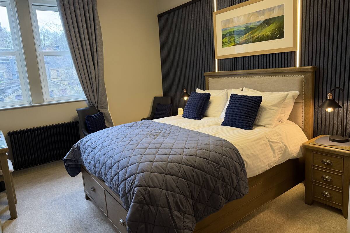 A neatly made bed with a quilted cover, a headboard with a framed landscape painting, a bedside lamp, and a view of the outside through a window with curtains.